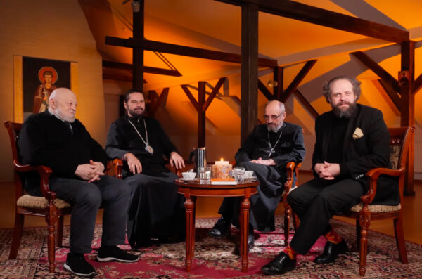 Czterech mężczyzn siedzi w fotelach wokół niewielkiego stolika z kawą i świecą, w ciepło oświetlonym, drewnianym wnętrzu. Scena ma charakter kameralnej rozmowy lub debaty; w tle widoczna jest ikona oraz elementy tradycyjnej architektury.