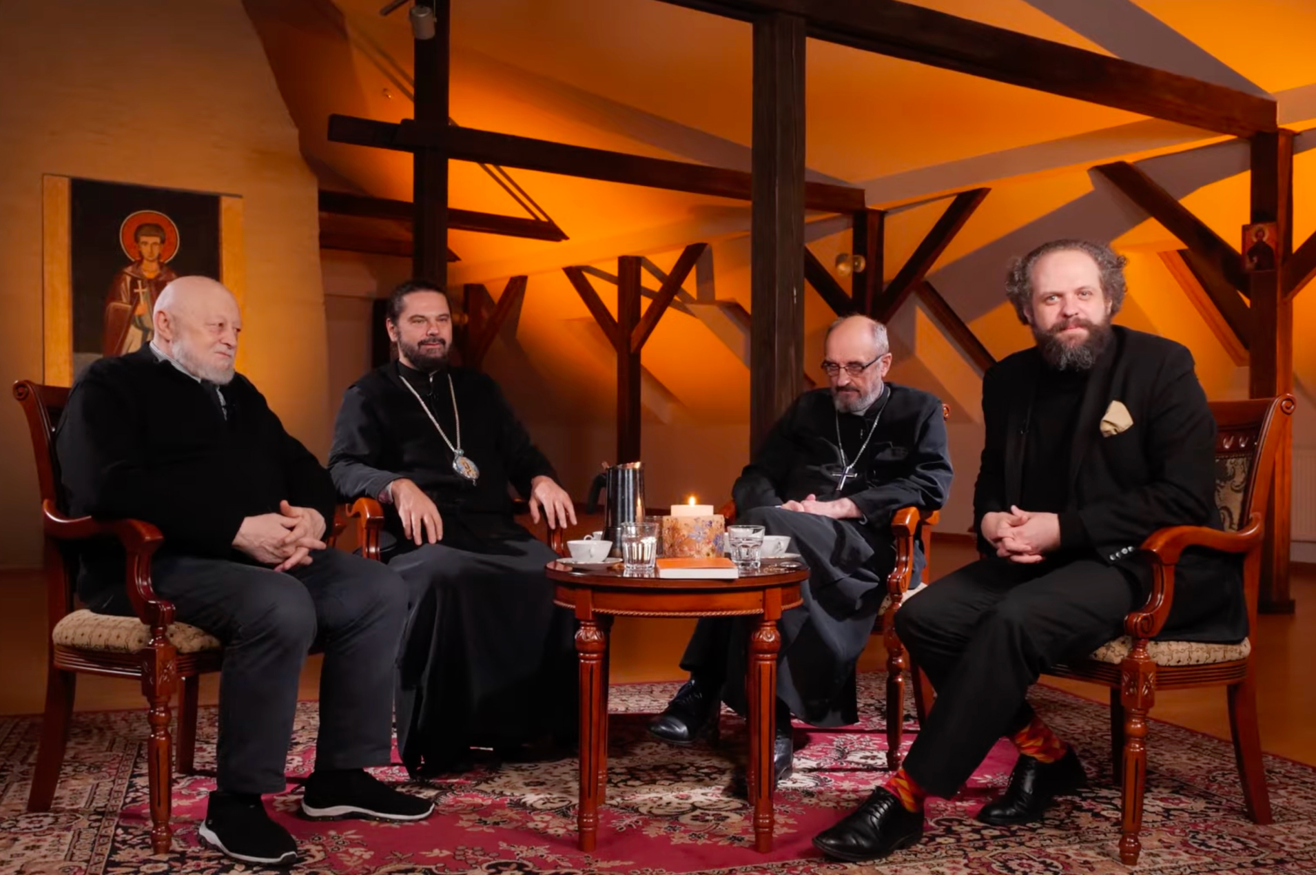 Czterech mężczyzn siedzi w fotelach wokół niewielkiego stolika z kawą i świecą, w ciepło oświetlonym, drewnianym wnętrzu. Scena ma charakter kameralnej rozmowy lub debaty; w tle widoczna jest ikona oraz elementy tradycyjnej architektury.